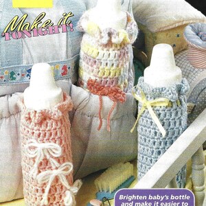 Könnte beinhalten: Drei gehäkelte Babyflaschen-Cozies in rosa, blau und mehrfarbigem Garn. Die Cozies sind mit Schleifen verziert und in einem Kinderbettchen zu sehen. Der Text "Make it TONIGHT!" ist im Bild sichtbar.