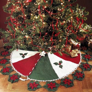 Può includere: Una gonna per albero di Natale fatta a mano in rosso, bianco e verde, con una ghirlanda di stelle di Natale all'uncinetto. La gonna è divisa in sezioni e presenta accenti di foglie di agrifoglio. L'albero è decorato con luci e nastri rossi.