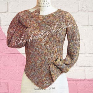 May include: A multi-colored knit sweater with long sleeves, displayed on a mannequin. The sweater features a unique, asymmetrical hemline and a textured knit pattern. The text "Chickie By the Sea" is written across the front.