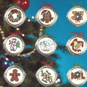 Peut inclure: Douze boules rondes en plastique transparent avec un tissu blanc &agrave; l'int&eacute;rieur, chacune pr&eacute;sentant un motif de point de croix sur le th&egrave;me de No&euml;l. Les motifs comprennent un flocon de neige, une couronne, un P&egrave;re No&euml;l, le mot "JOIE", un chien, un ange, un ours polaire, une colombe avec le mot "PAIX", une cloche, un loup, un bonhomme en pain d'&eacute;pices, l'ann&eacute;e "1985" et le mot "NOEL".