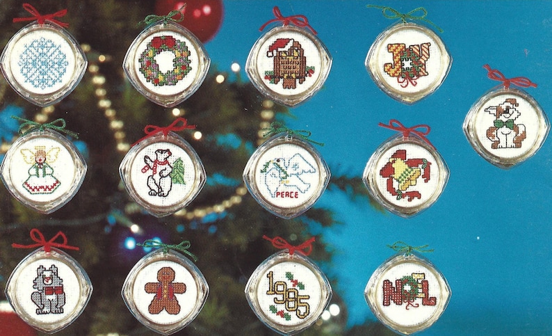 Peut inclure: Douze boules rondes en plastique transparent avec un tissu blanc &agrave; l'int&eacute;rieur, chacune pr&eacute;sentant un motif de point de croix sur le th&egrave;me de No&euml;l. Les motifs comprennent un flocon de neige, une couronne, un P&egrave;re No&euml;l, le mot "JOIE", un chien, un ange, un ours polaire, une colombe avec le mot "PAIX", une cloche, un loup, un bonhomme en pain d'&eacute;pices, l'ann&eacute;e "1985" et le mot "NOEL".