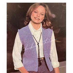 Może przedstawiać: Purpurowy, dzianinowy kamizelka z dekoltem w kształcie litery V i czarnym krawatem bolo. Kamizelka jest noszona na białej koszuli z guzikami i szarych spodniach.