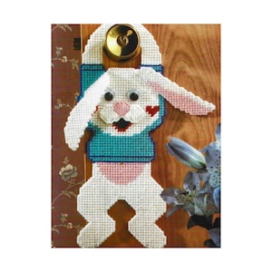 Puede incluir: Un colgador de puerta de conejo blanco con detalles azules y rosas. El conejo está agarrado a un pomo de puerta con sus patas delanteras y tiene un corazón en la mejilla.