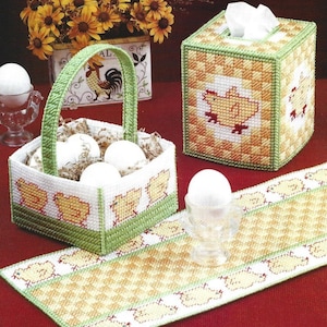 Pode incluir: Um conjunto de três itens de crochê com tema de Páscoa: uma capa para caixa de lenços, uma cesta com ovos e um corredor de mesa. Os três itens apresentam um padrão xadrez com quadrados amarelos e brancos e uma borda verde e branca. A capa da caixa de lenços e o corredor de mesa também apresentam um padrão repetido de pintinhos amarelos.