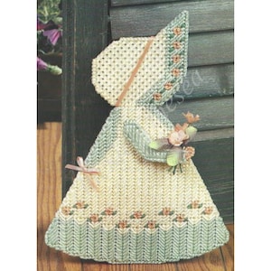 May include: A crocheted doll wearing a white dress with a green and white trim, a white bonnet with a pink ribbon, and holding a bouquet of pink flowers.