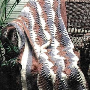 May include: A handmade crochet blanket draped over a wicker chair. The blanket features a repeating chevron pattern in shades of brown, white, and gray. The texture of the blanket is visible, with a soft, cozy appearance. The chair is a dark brown color.