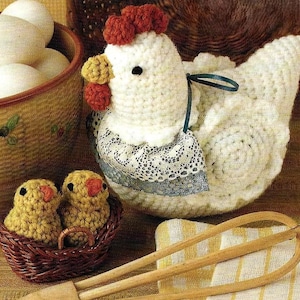 May include: A crocheted white hen with a red comb and a blue ribbon around its neck. The hen is sitting on a table with two small crocheted chicks in a brown wicker basket. There is a wooden whisk and a white and yellow striped towel in the background.