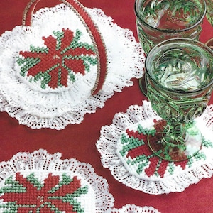 Puede incluir: Tres posavasos de ganchillo blanco con diseños de poinsettia rojos y verdes. Los posavasos están colocados sobre una superficie roja con dos copas de vidrio verde.