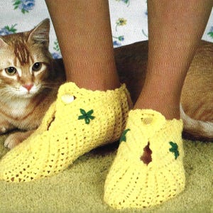 May include: A pair of yellow crochet slippers with a green flower design and a button closure. The slippers are worn on a person's feet. A cat is in the background.
