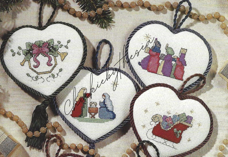 May include: Four heart-shaped Christmas ornaments with cross-stitch designs. The ornaments feature a nativity scene, a sleigh with presents, a star, and a bell with a bow.