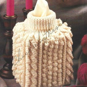 May include: A crocheted tissue box cover in a cream color with a textured design. The cover is sitting on a gray surface with a pink yarn ball and crochet hook.
