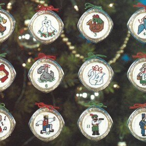 Peut inclure: Douze boules de No&euml;l rondes en verre transparent avec un fond blanc et des motifs de point de croix color&eacute;s. Chaque boule est orn&eacute;e d'un ruban rouge et d'un num&eacute;ro de 1 &agrave; 12.