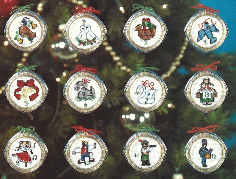 Peut inclure: Douze boules de No&euml;l rondes en verre transparent avec un fond blanc et des motifs de point de croix color&eacute;s. Chaque boule est orn&eacute;e d'un ruban rouge et d'un num&eacute;ro de 1 &agrave; 12.