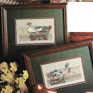 May include: Two framed cross-stitch embroidery pieces depicting ducks resting on a wooden surface. The top piece features a red-headed duck, while the bottom piece features a mallard duck.