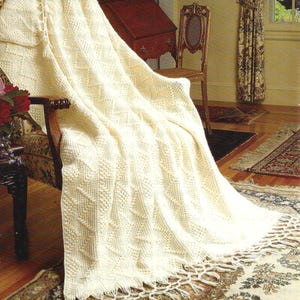 May include: Cream-colored knitted throw blanket with a textured pattern and fringe detail. The blanket is draped over a patterned armchair, showcasing its intricate design and cozy appeal. The throw blanket is a home decor item.