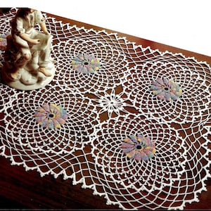 May include: A white, crocheted table runner with a repeating circular pattern. The runner features colorful, flower-shaped accents and a scalloped edge. A decorative figurine is placed on the runner.