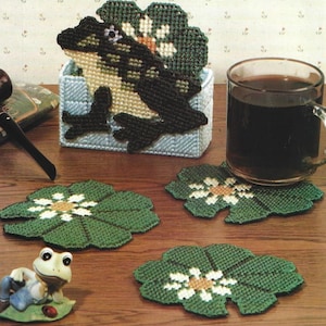 May include: A set of four green and white crocheted lily pad coasters with a frog sitting on a lily pad and a frog-shaped tissue box holder. The tissue box holder has a green and white crocheted frog design.