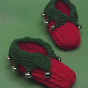 May include: A pair of handmade red crochet slippers with green trim and silver bells. The slippers have a festive, elf-like design, suitable for the holiday season. The slippers are made of soft yarn.