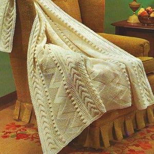 May include: A white knitted throw blanket with a cable knit pattern is draped over a chair. The blanket has a textured, woven design and is made of soft yarn.