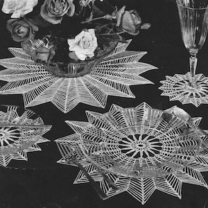 May include: A set of white crocheted star-shaped placemats and coasters featuring intricate radial patterns. The set includes a large centerpiece mat under a glass bowl of roses, a plate mat with silverware, and a small coaster under a glass goblet.