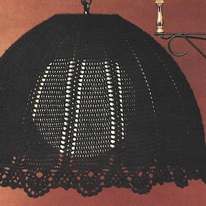 May include: A black, crocheted lampshade with a scalloped edge and an openwork design. The lampshade is suspended from a metal hook and has a semi-circular shape. The design features vertical lines and a circular pattern.
