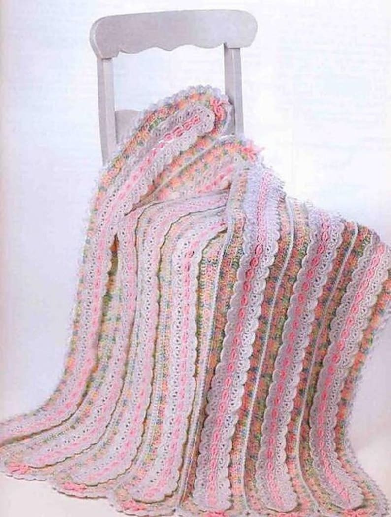 Puede incluir: Una manta de beb&eacute; de ganchillo con un delicado borde de encaje en tonos de rosa, blanco y verde. La manta est&aacute; drapeada sobre una silla blanca.