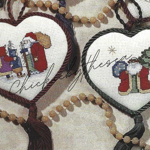 May include: Two white heart-shaped ornaments with red and green trim. One ornament features a Santa Claus figure with two children. The other ornament features a Santa Claus figure with a Christmas tree.