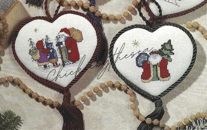 May include: Two white heart-shaped ornaments with red and green trim. One ornament features a Santa Claus figure with two children. The other ornament features a Santa Claus figure with a Christmas tree.