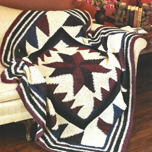 May include: A crocheted throw blanket with a geometric pattern in shades of white, burgundy, navy blue, and green. The blanket is draped over a cream-coloured sofa.