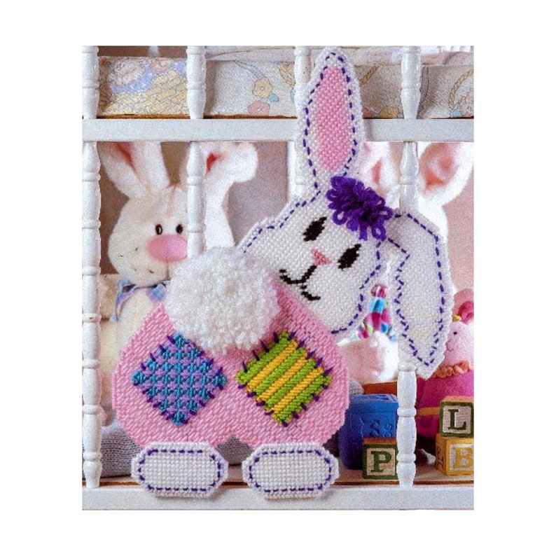 May include: A pink and white bunny with a purple flower on its head. The bunny is made of plastic canvas and has a patchwork design on its body. The bunny is hanging on a white crib.