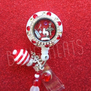 May include: A white and red striped charm hangs from a white retractable badge holder with a clear plastic badge holder. The badge holder has a round, clear, glass locket with a white background and red rhinestones around the edge. Inside the locket are various charms, including a red blood drop, a microscope, a caduceus, and a small book with the word "Justice" on it.