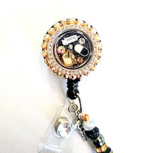 May include: A gold and black retractable badge holder with a clear round charm featuring a syringe, pill, and other medical symbols. The charm is surrounded by rhinestones. The holder has a black beaded chain with a clear crystal charm.