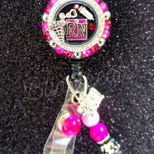 May include: A black and gold retractable badge holder with a pink and gold charm. The charm has a pink background with the letters "RN" in gold and a medical symbol. The badge holder has pink and gold beads.