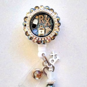 May include: A round, white retractable badge reel with a decorative face featuring the letters "RX", a pill, and a medicine bottle. The reel is surrounded by a ring of iridescent rhinestones and has a charm with a cross and heart.