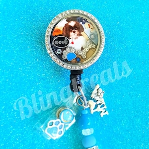 May include: A silver charm with a clear glass locket featuring a photo of a dog with the word "woof" inside. The charm is attached to a blue and silver beaded chain with a paw print charm and a small dog charm.