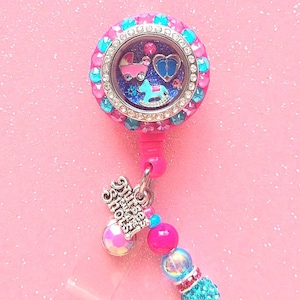 May include: A pink and blue jeweled charm with a clear glass locket containing a baby carriage, rocking horse, and a heart. The charm is attached to a pink and blue beaded lanyard with a silver charm that says "It's a Girl Shower".