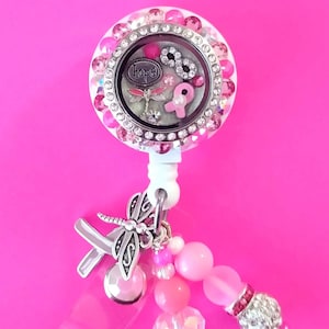 May include: A pink and silver charm with a clear glass locket featuring a pink ribbon, a dragonfly, and the word "hope". The charm is attached to a white retractable badge holder with a pink and silver beaded chain.