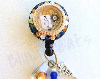 May include: A blue and gold eye chart themed retractable badge holder with a clear charm containing an eye chart, a pair of glasses, a blue eye, and a silver medical symbol. The badge holder has a blue and gold beaded chain with a silver charm.