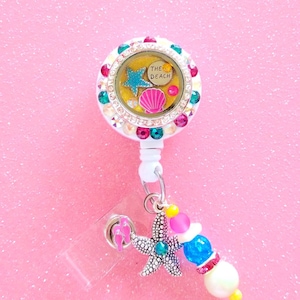 May include: A white and rhinestone embellished badge reel with a beach theme. The reel features a clear, round center with a blue starfish, pink seashell, and the words "THE BEACH". A starfish charm and beaded accents dangle below.