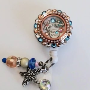 May include: A white and gold retractable badge holder with a clear charm that has a starfish, flip flops, and the text "THE BEACH" inside. The charm is surrounded by rhinestones. The badge holder has a silver starfish charm and a clear rhinestone charm attached to it.