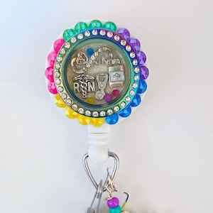 May include: A rainbow-colored retractable badge holder with a clear charm featuring a stethoscope, a heart, an infinity symbol, a bottle of pills, and the letters "RN". The charm is surrounded by rhinestones. A white angel charm with a yellow bead and purple and turquoise beads hangs from the badge holder.