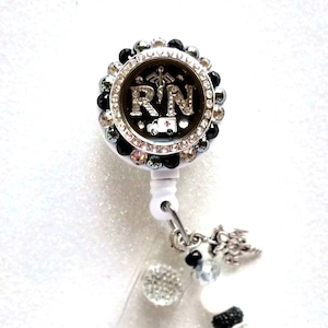 May include: A black and silver rhinestone badge reel with a black and white 'RN' charm and a silver medical symbol. The charm is surrounded by rhinestones and has a small red ambulance on it. The reel has a white plastic clip and a black and white beaded chain.