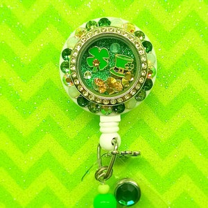 May include: A white retractable badge holder with a green, gold, and white glitter filled charm featuring a shamrock and a leprechaun hat. The charm is surrounded by clear rhinestones and green crystals. The badge holder has a green and white beaded chain.