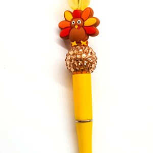 May include: A yellow pen with a brown and orange turkey charm and a gold rhinestone ball at the top.