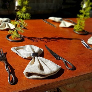 May include: A table setting with a rustic, hand-forged cutlery set. The set includes a knife, fork, and napkin ring. The napkin is folded into a bow shape and placed on the table. The cutlery is made of metal and has a twisted design.