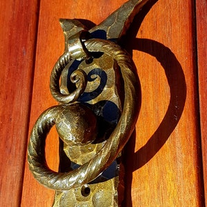 May include: A gold metal door knocker shaped like a crescent moon with a twisted rope design and a round ball at the end. The knocker is mounted on a wooden door.
