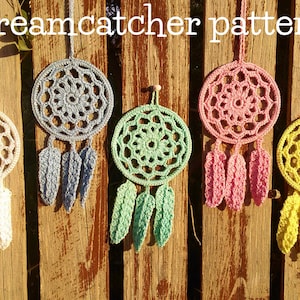 Pode incluir: Cinco apanhadores de sonhos de crochê pendurados em uma cerca de madeira. Os apanhadores de sonhos são de várias cores, incluindo branco, azul, verde, rosa e amarelo. Cada apanhador de sonhos tem um design de teia circular e penas penduradas na parte inferior.