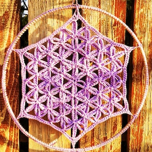 以下が含まれることがあります： 手作りのドリームキャッチャー。円形のフレームと中心の幾何学模様が特徴です。複雑な模様はラベンダーと紫の色合いでかぎ針編みされており、繊細で芸術的な作品です。ドリームキャッチャーは木製の背景に吊るされています。