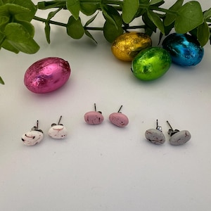 May include: Three pairs of small, oval-shaped earrings with different coloured patterns. The earrings are white with pink accents, pink with white accents, and grey with white accents. The earrings are displayed on a white surface.