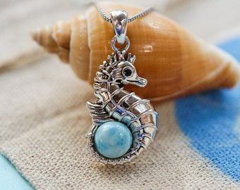 May include: Silver seahorse pendant with a blue and white gemstone. The pendant is hanging from a silver chain and is resting on a seashell.